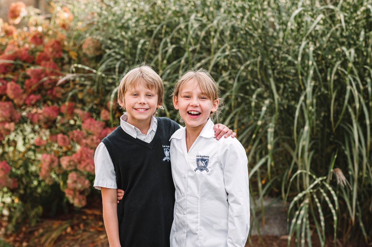 Children in matching school uniforms pose together in front of seasonal red and green landscaping.