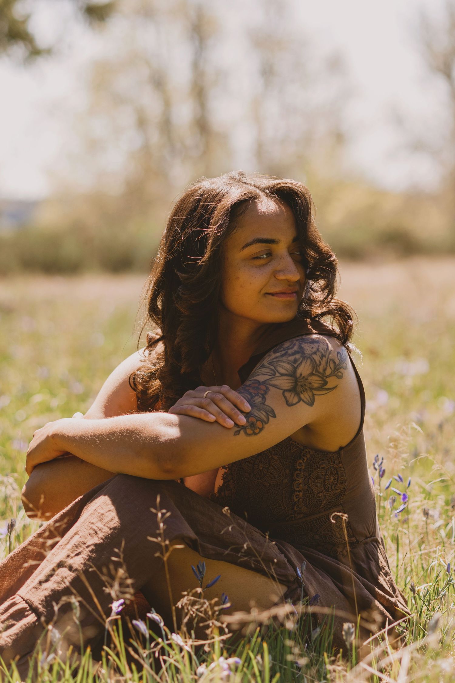 A young woman with a floral arm tattoo sits cross-legged in a sunny meadow, smiling softly in a brown dress.