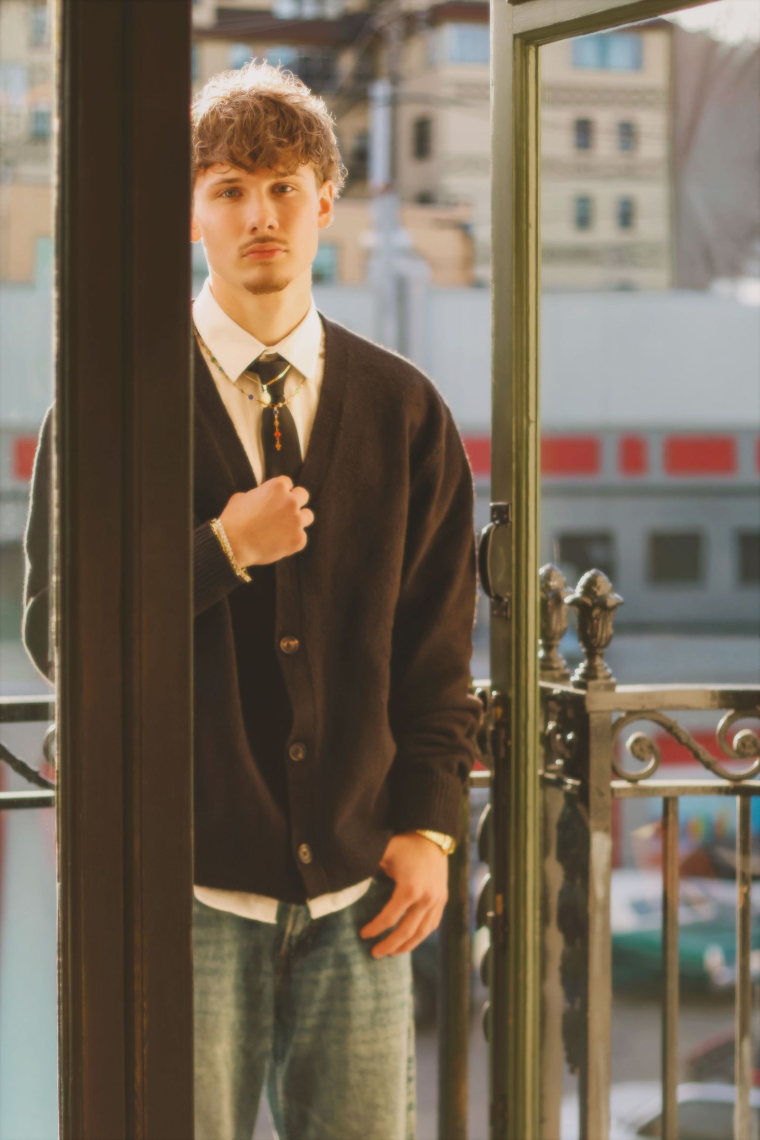 A person in a black cardigan, white shirt and tie stands by a glass door in warm lighting.