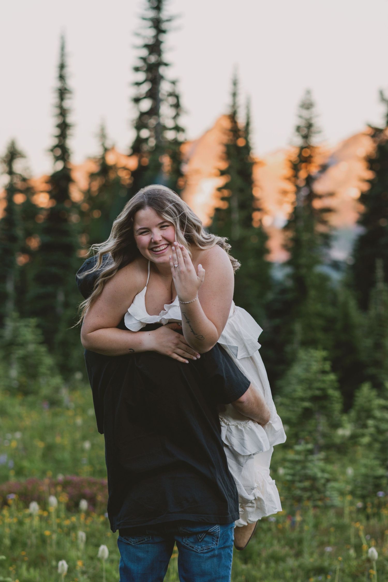 Joyful woman shows engagement ring while being embraced by partner in a mountain meadow at golden hour.