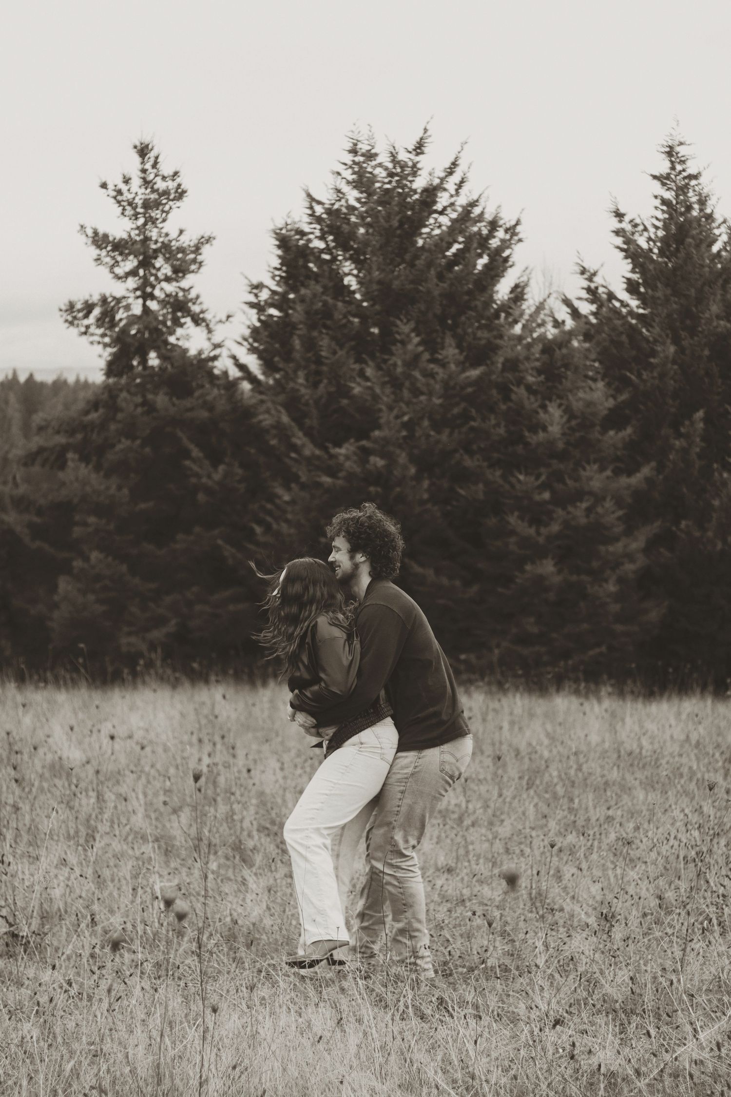 A couple embraces in a field with evergreen trees in the background, captured in black and white photography.