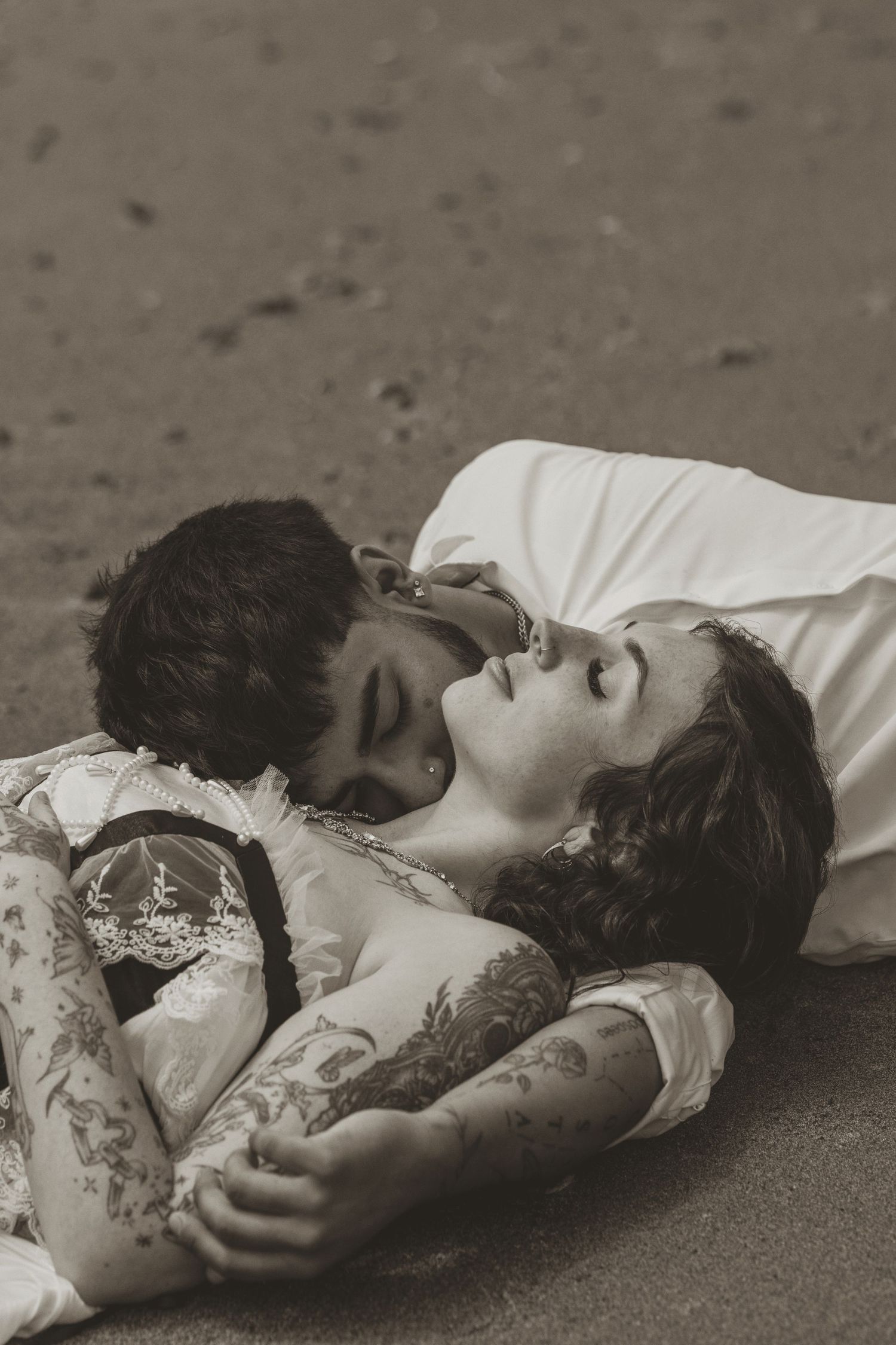 Black and white photo of a tattooed couple lying on sand, sharing a tender romantic moment together.