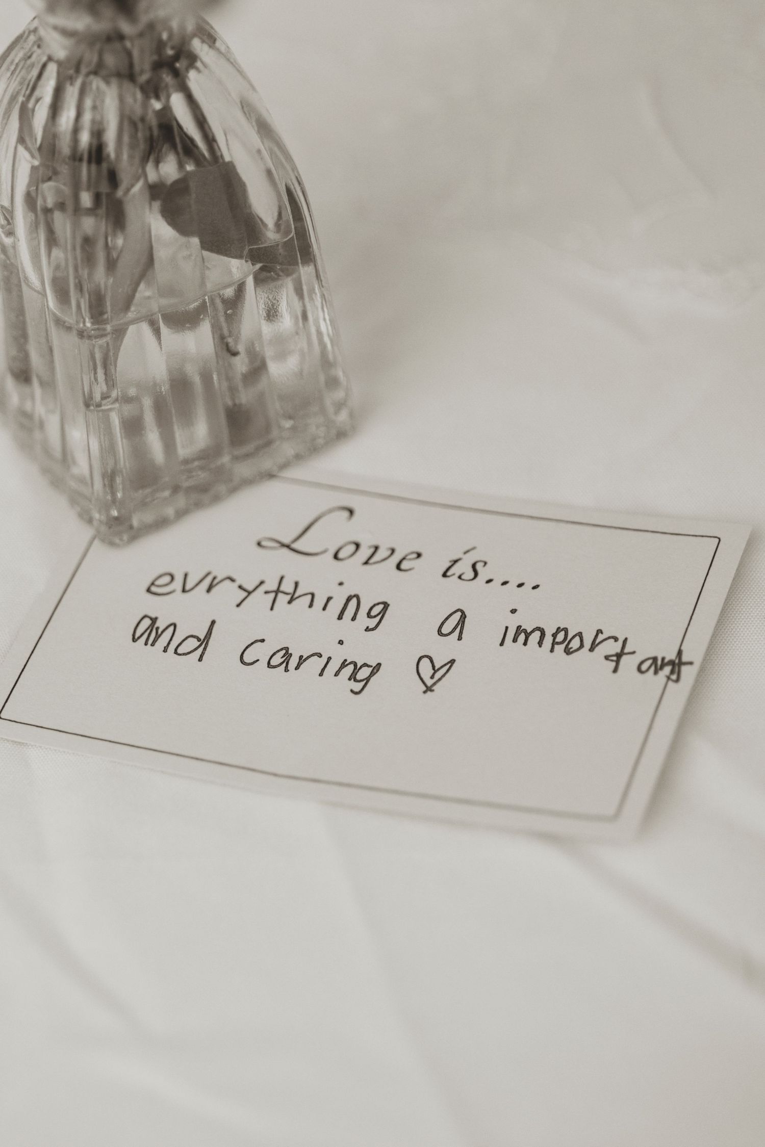 Handwritten note reading Love is everything important and caring with a heart, beside a glass perfume bottle on white fabric.