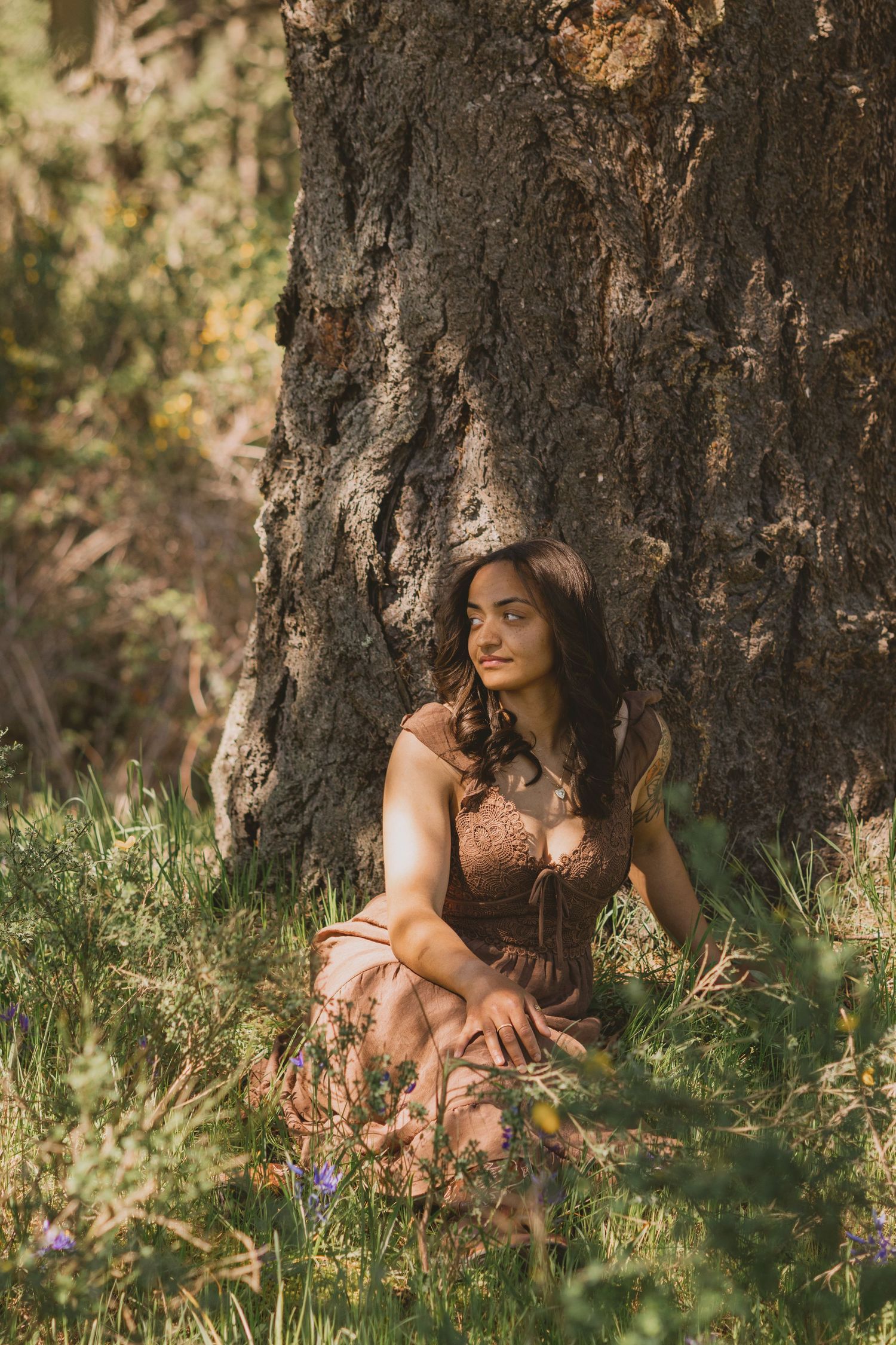 Young woman in brown dress sitting against large tree trunk in sunlit forest with wildflowers.