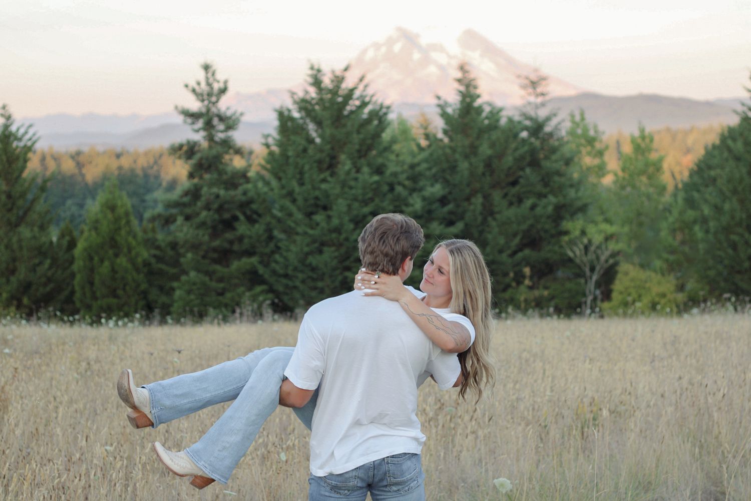 A romantic sunset photoshoot in a golden wheat field with mountains and evergreen trees in the background.