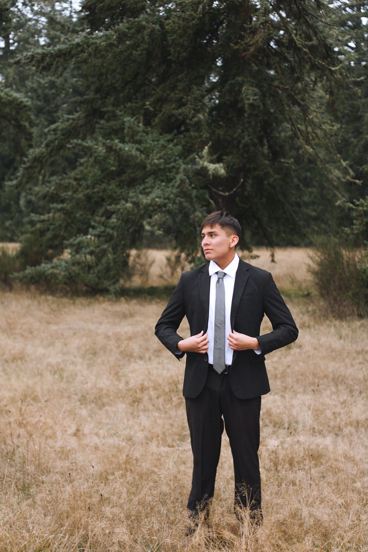 Person in black suit and white tie stands in brown grass field with evergreen trees in background.