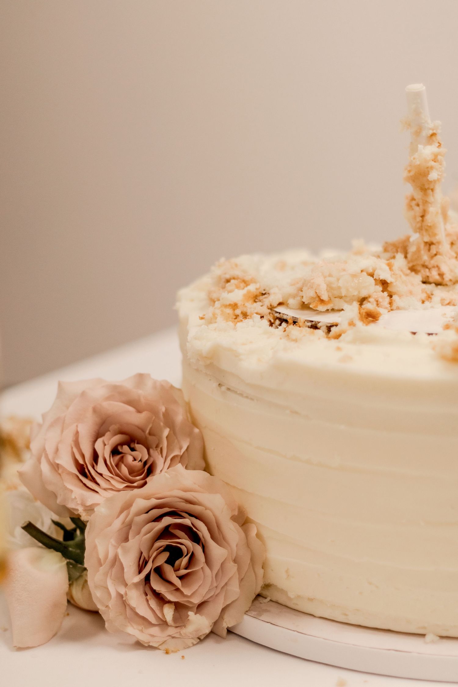 Elegant white frosted cake topped with crumbles and a candle, surrounded by dusty pink roses on a white surface.