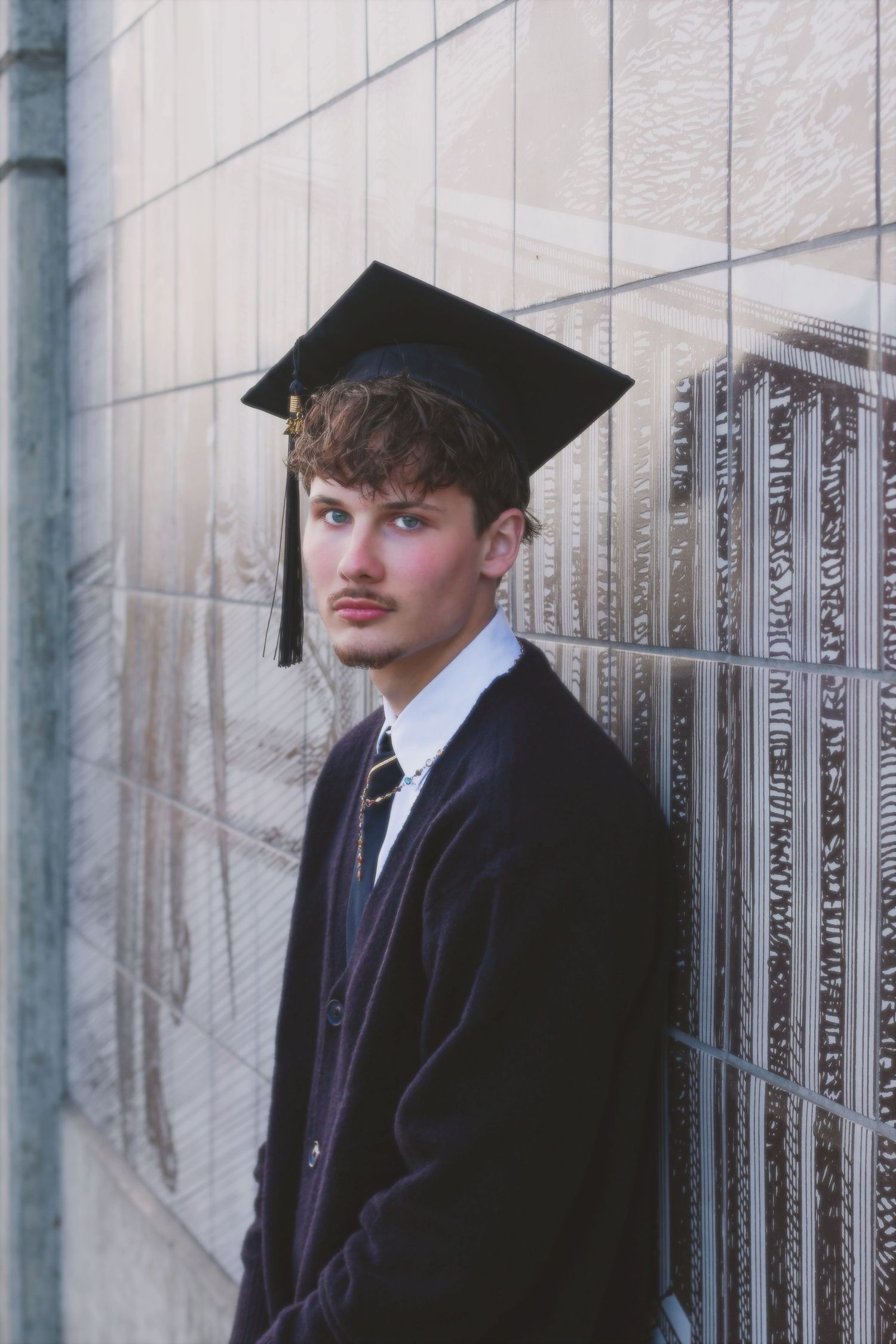 Two graduates in black caps and gowns stand against a white wall with decorative columns.