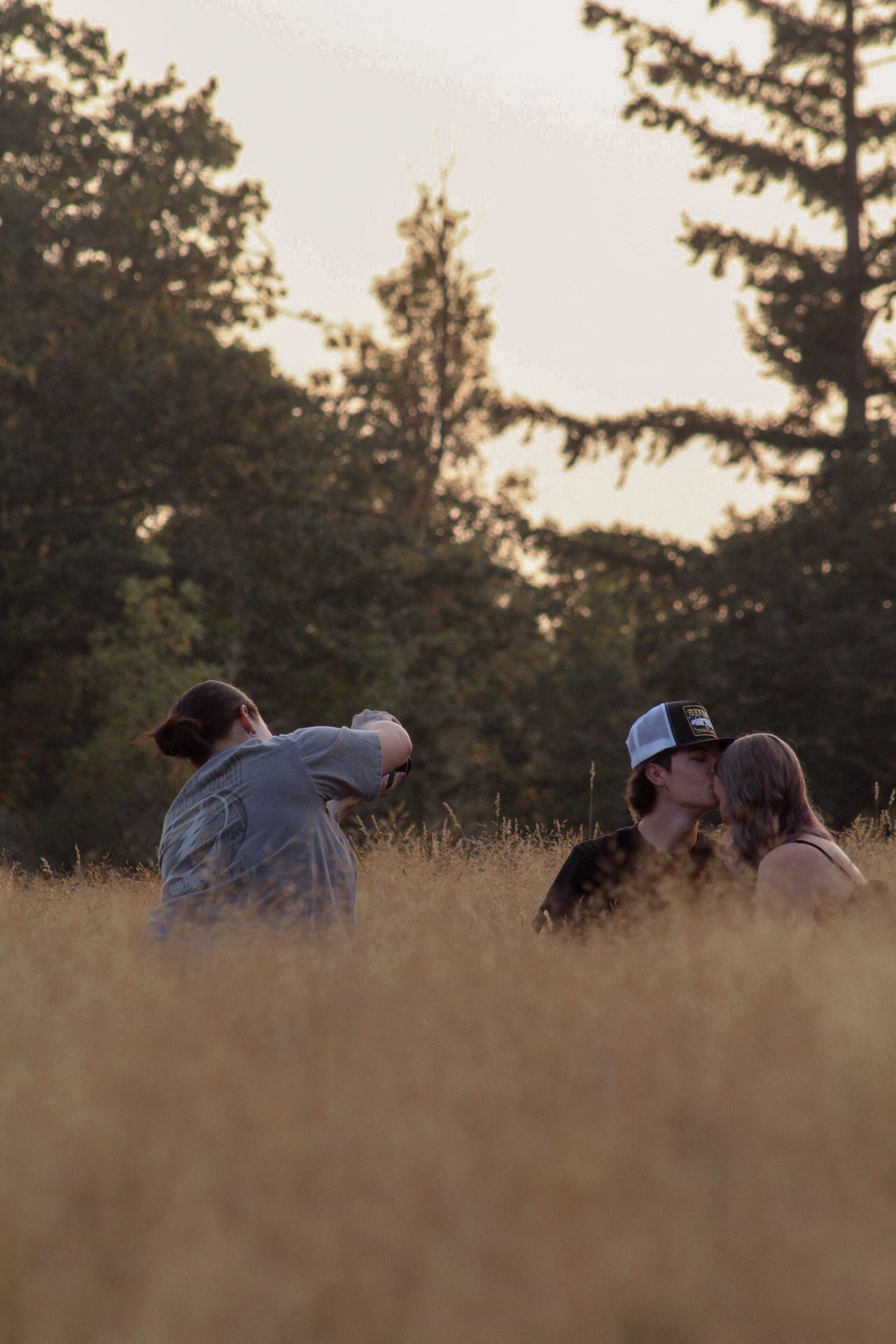A couple lying in tall grass during golden hour with evergreen trees silhouetted against the sky.