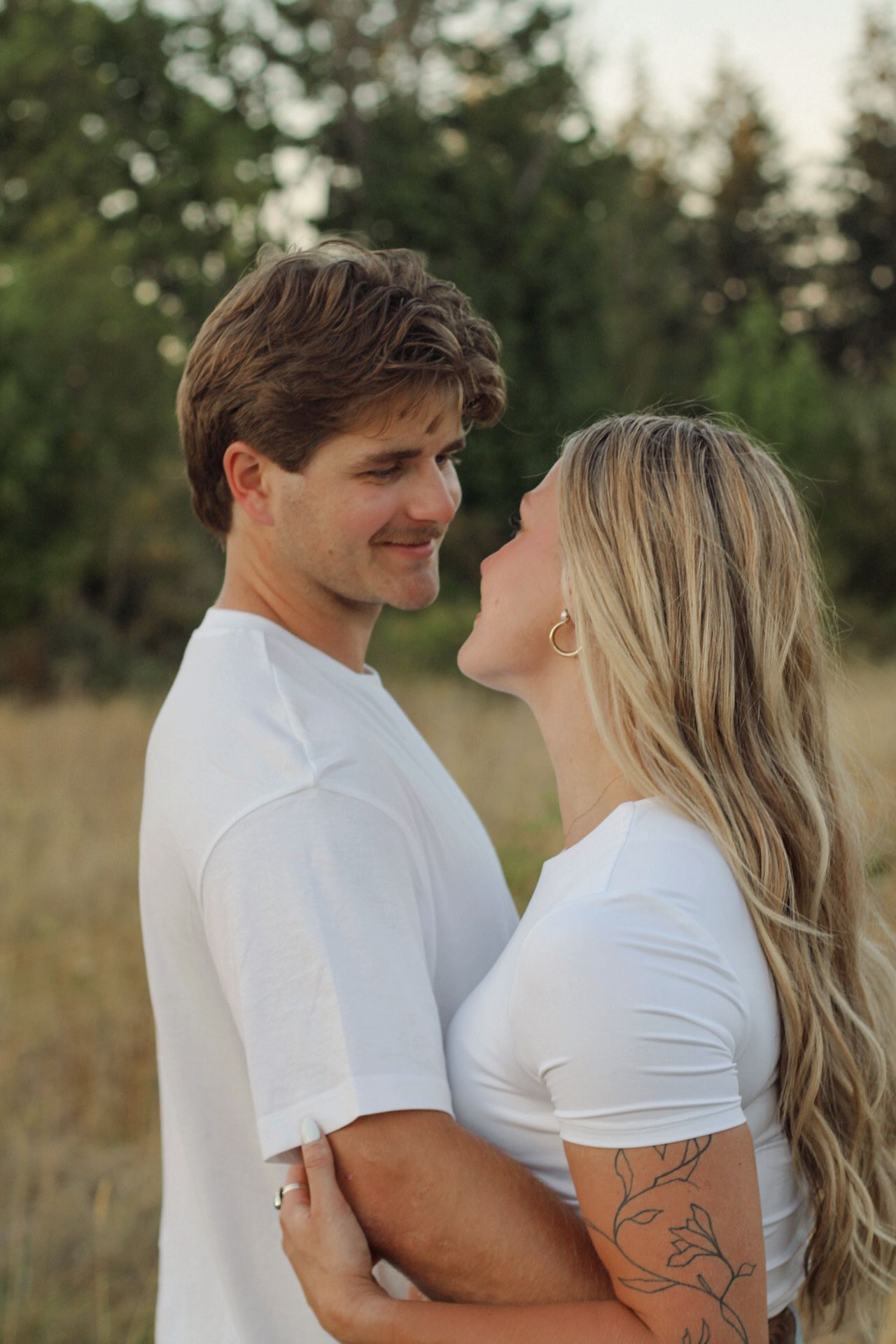 Couple in white shirts embracing outdoors in a field with trees in the background.