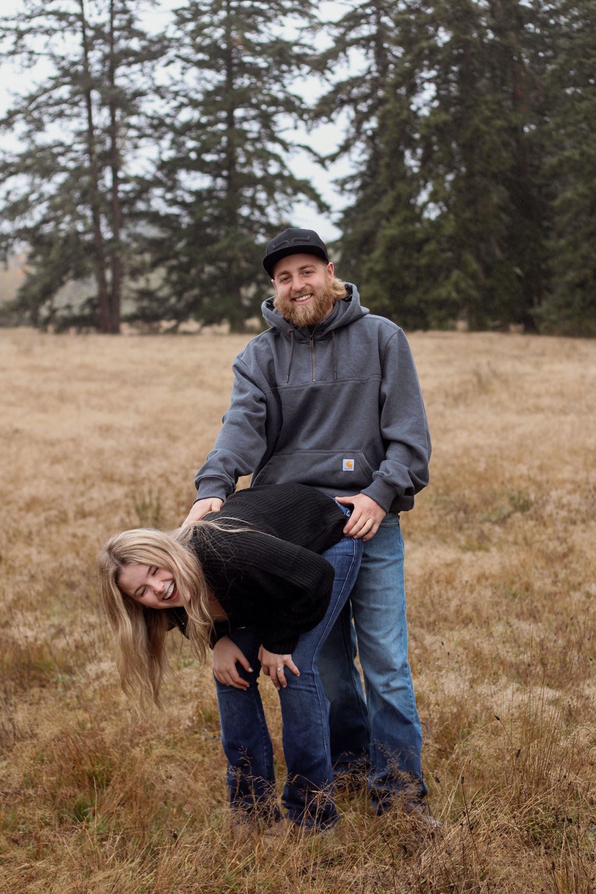 A couple shares a playful moment together while sitting in a golden field of dry grass with evergreen trees in the background.