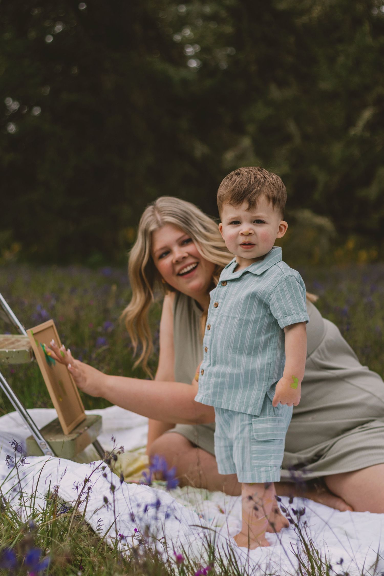 A smiling woman sits in a wildflower field with a toddler boy in a green outfit on a white blanket outdoors.