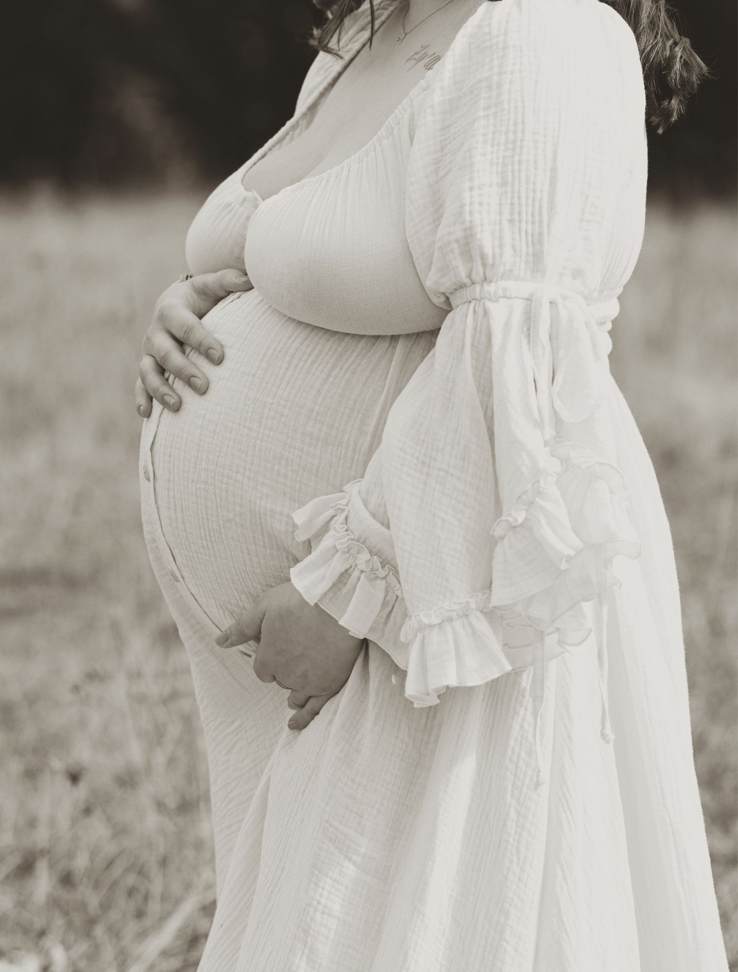 Pregnant woman in white dress cradling baby bump outdoors in sepia-toned maternity photo.