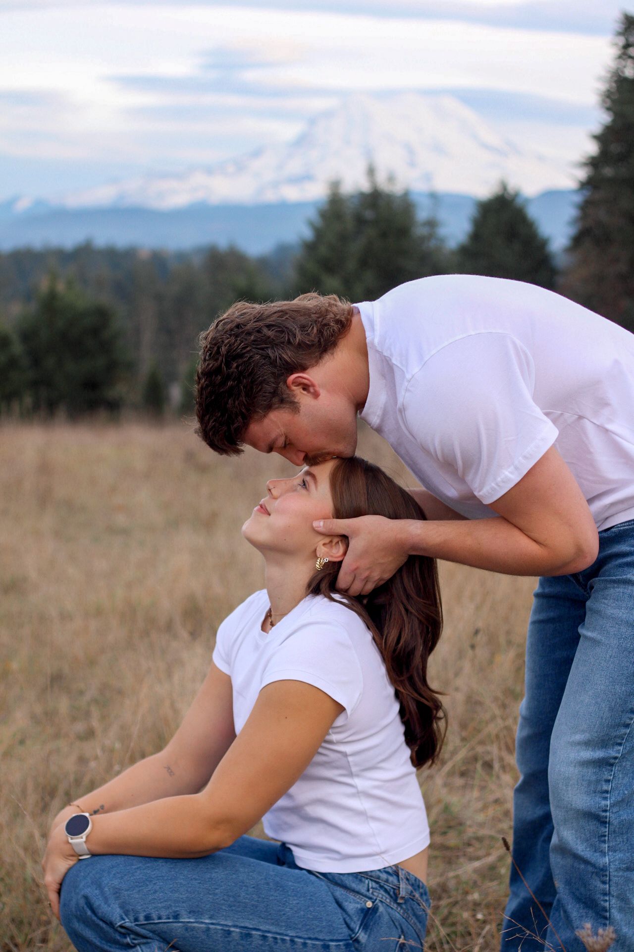 A couple in white shirts and jeans embrace lovingly in a grassy field with mountains visible in the background.