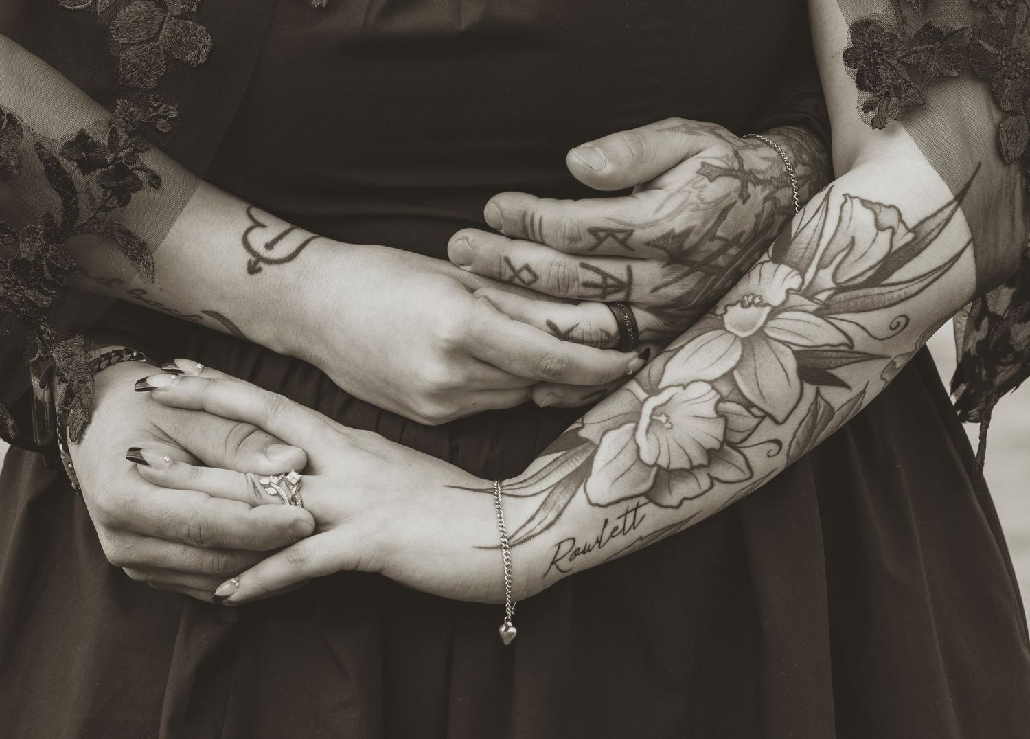Black and white photo of tattooed arms with floral and fine line tattoos, hands clasped together elegantly.