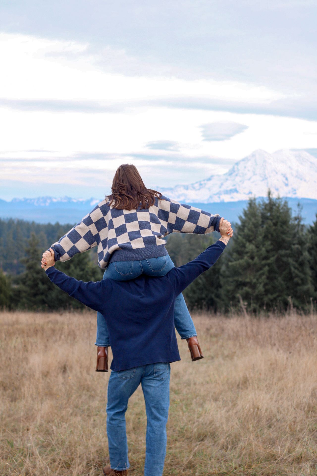 Two people enjoy a scenic mountain view from a grassy field on a cloudy day while one sits on the other's shoulders.