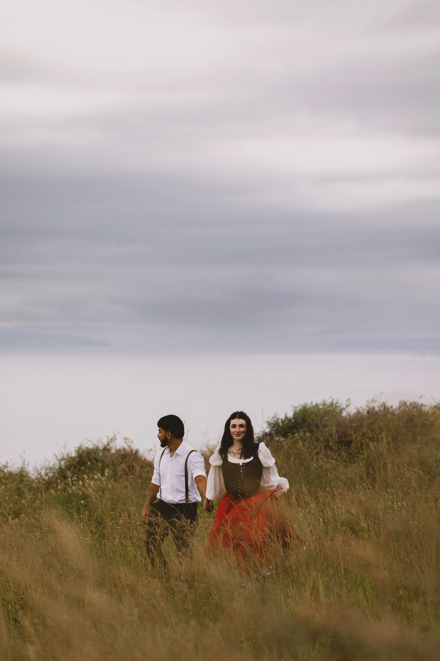 Couple in vintage attire walking through tall grass field under cloudy sky, woman in red skirt and man in suspenders.