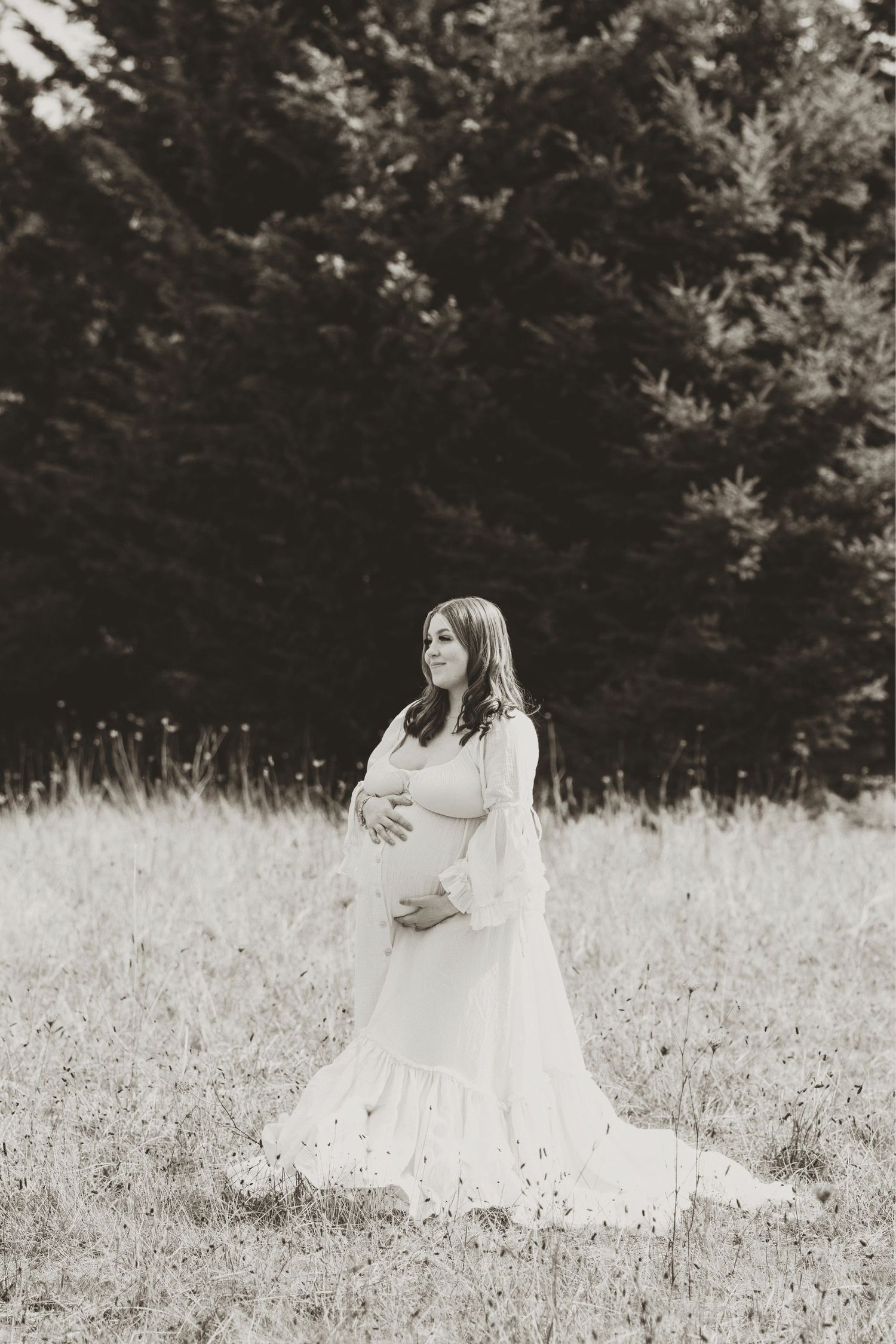 Pregnant woman in white dress standing in field with evergreen trees in black and white maternity photo.