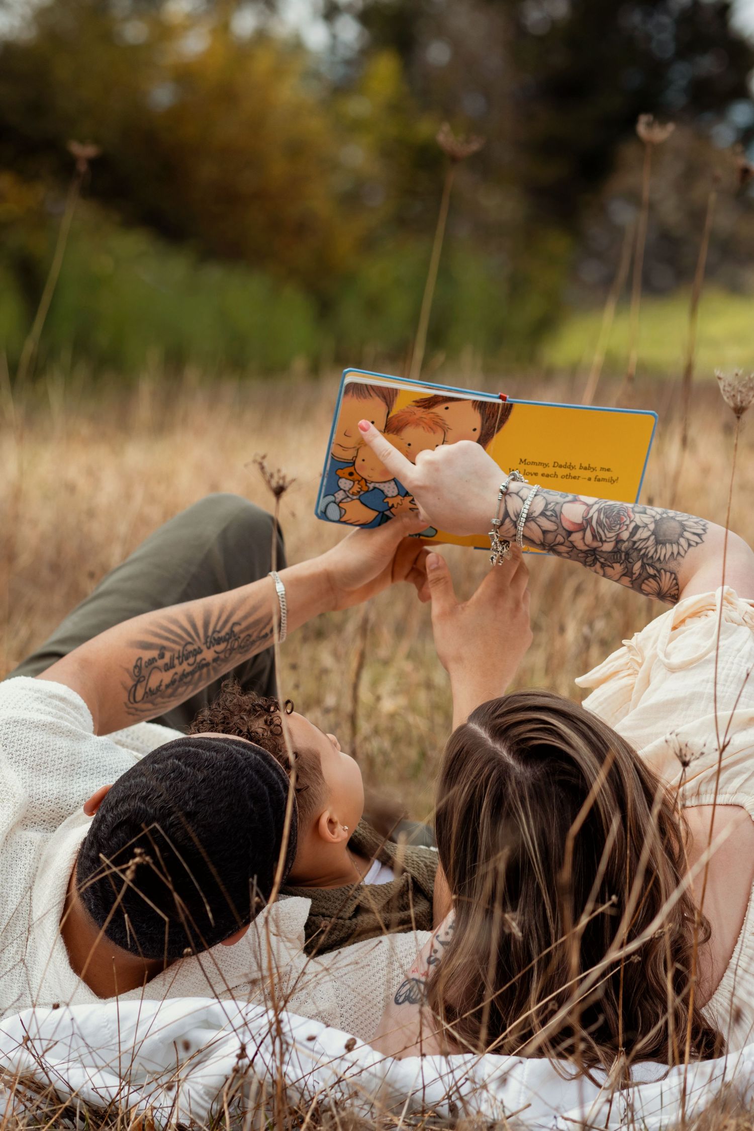 Tattooed couple lying in a field of tall grass, reading a yellow children's book together outdoors.