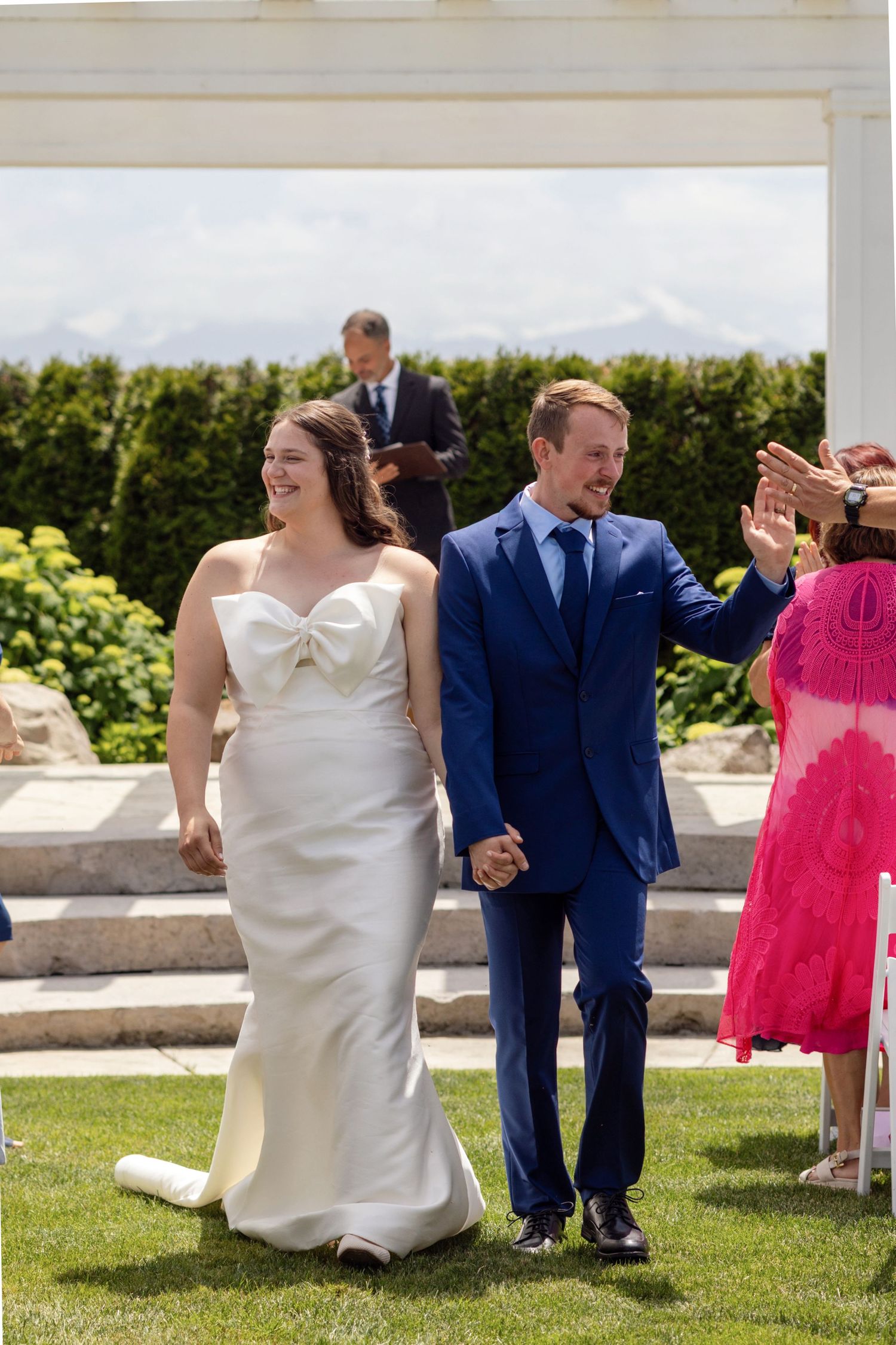 Bride in white gown and groom in blue suit walk hand-in-hand after outdoor wedding ceremony on sunny day.