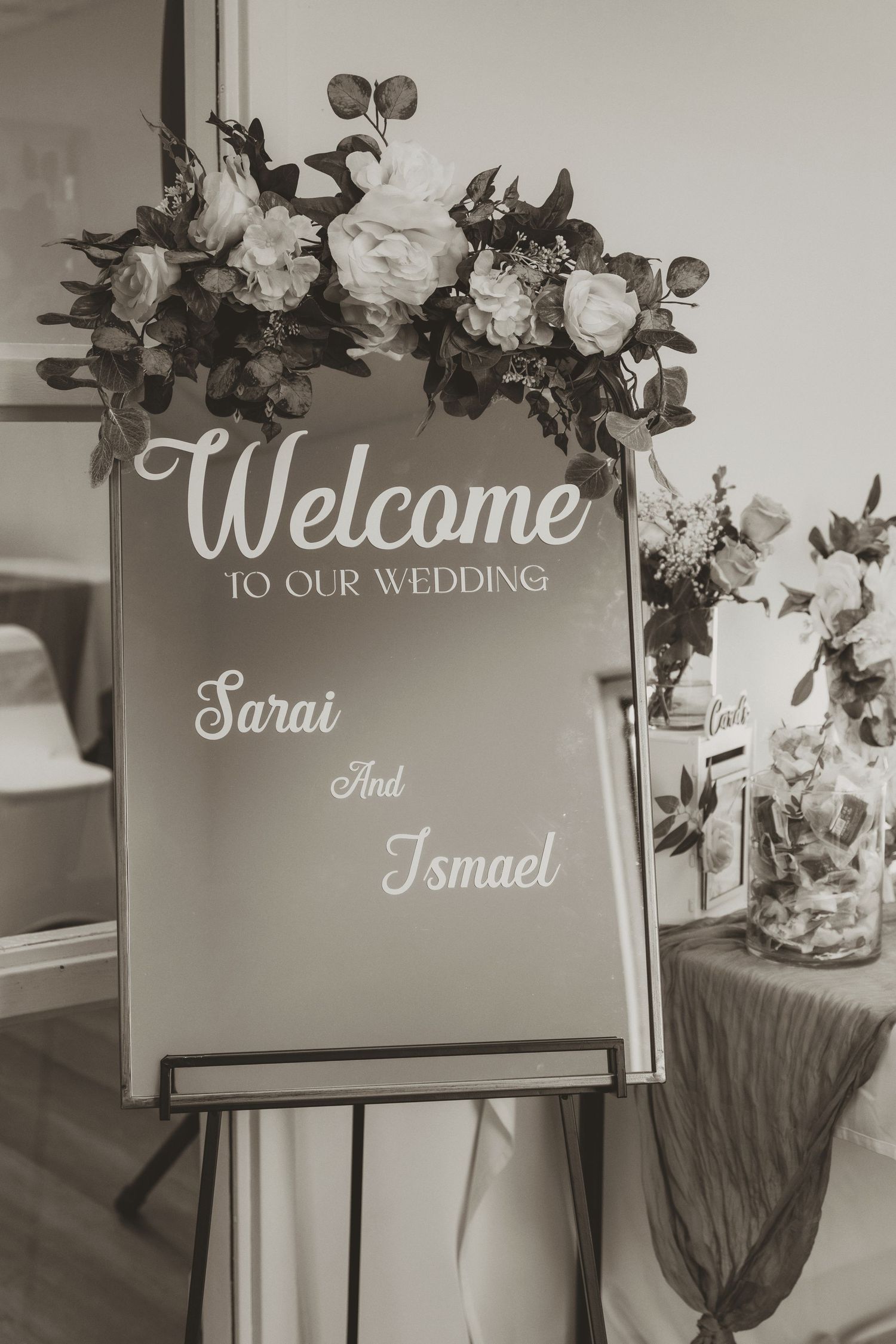 Black and white wedding welcome sign for Sarai and Ismael decorated with white roses and greenery on an easel.
