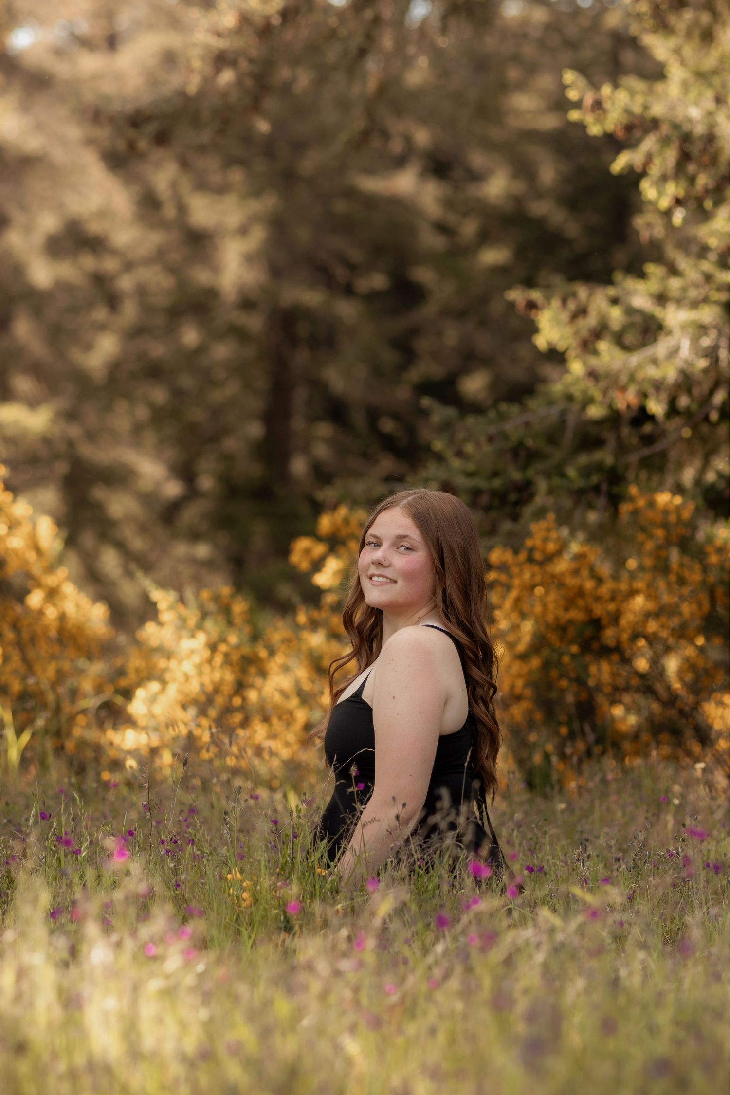 Young woman in black dress smiling in a wildflower meadow with golden autumn trees in background.