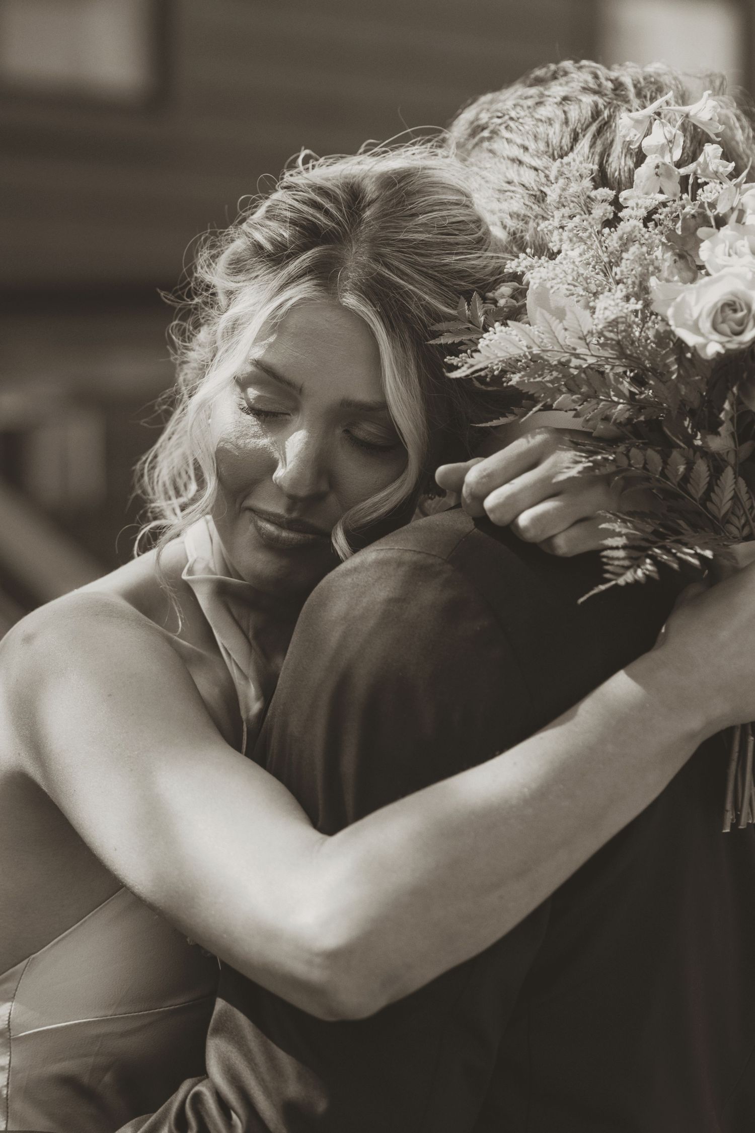 Black and white photo of a bride embracing someone while holding a floral bouquet, eyes closed with emotion.