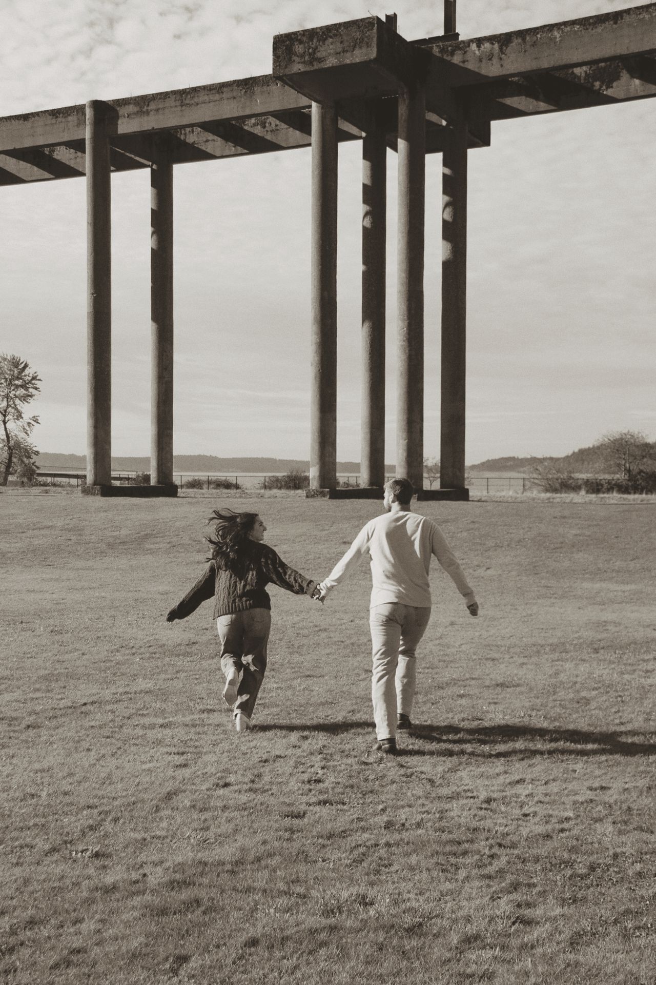 Two people walk and play together beneath large architectural pillars in a grassy field on an overcast day.