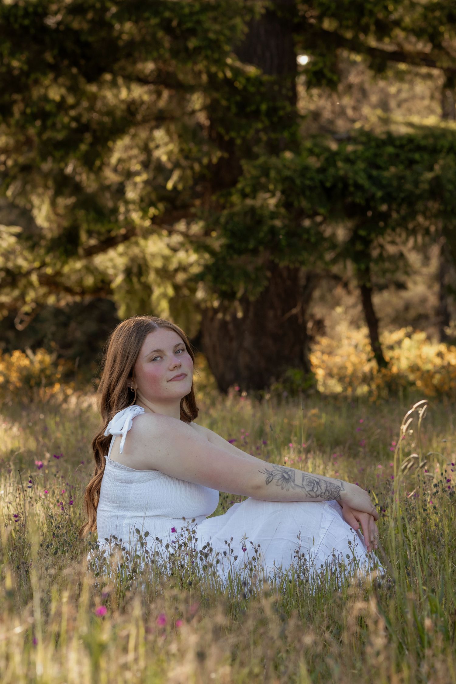 Woman in white dress sitting in wildflower meadow with golden sunlight and green trees in background.
