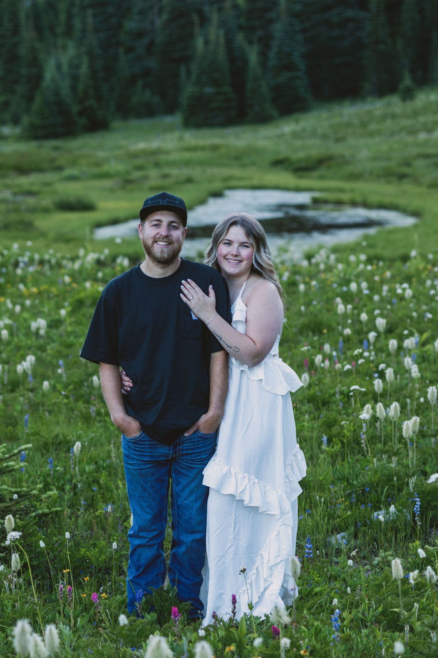 Couple posing in a wildflower meadow, woman in white dress and man in black shirt and jeans, with a stream and forest behind.
