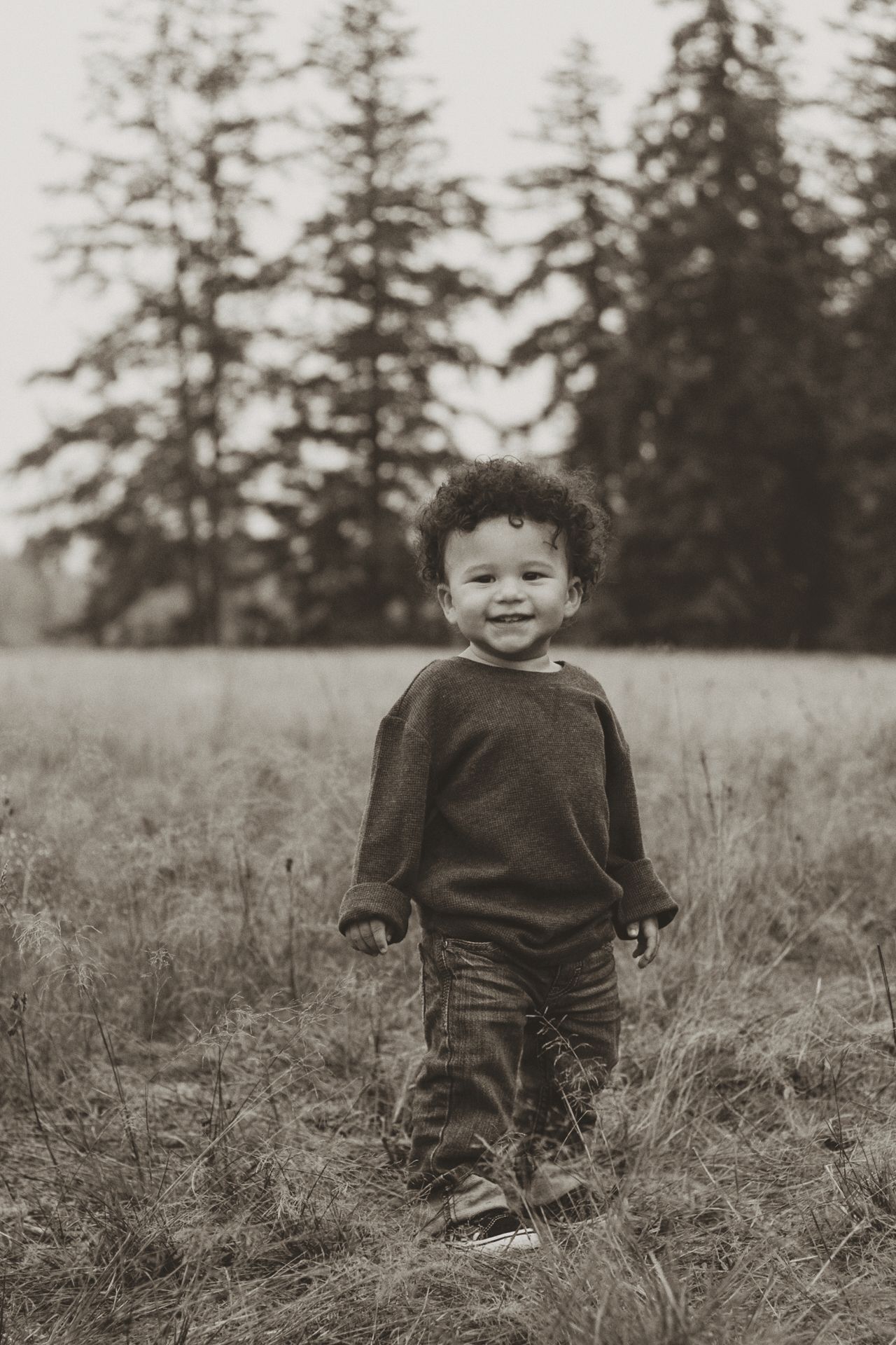 A small child stands in a grassy field with tall evergreen trees in the background, captured in black and white.