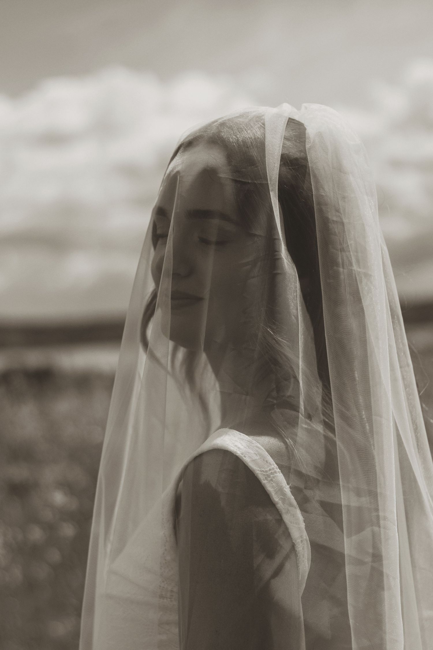 Serene bride in sepia tone with sheer veil draped over head, eyes closed, against cloudy sky backdrop.
