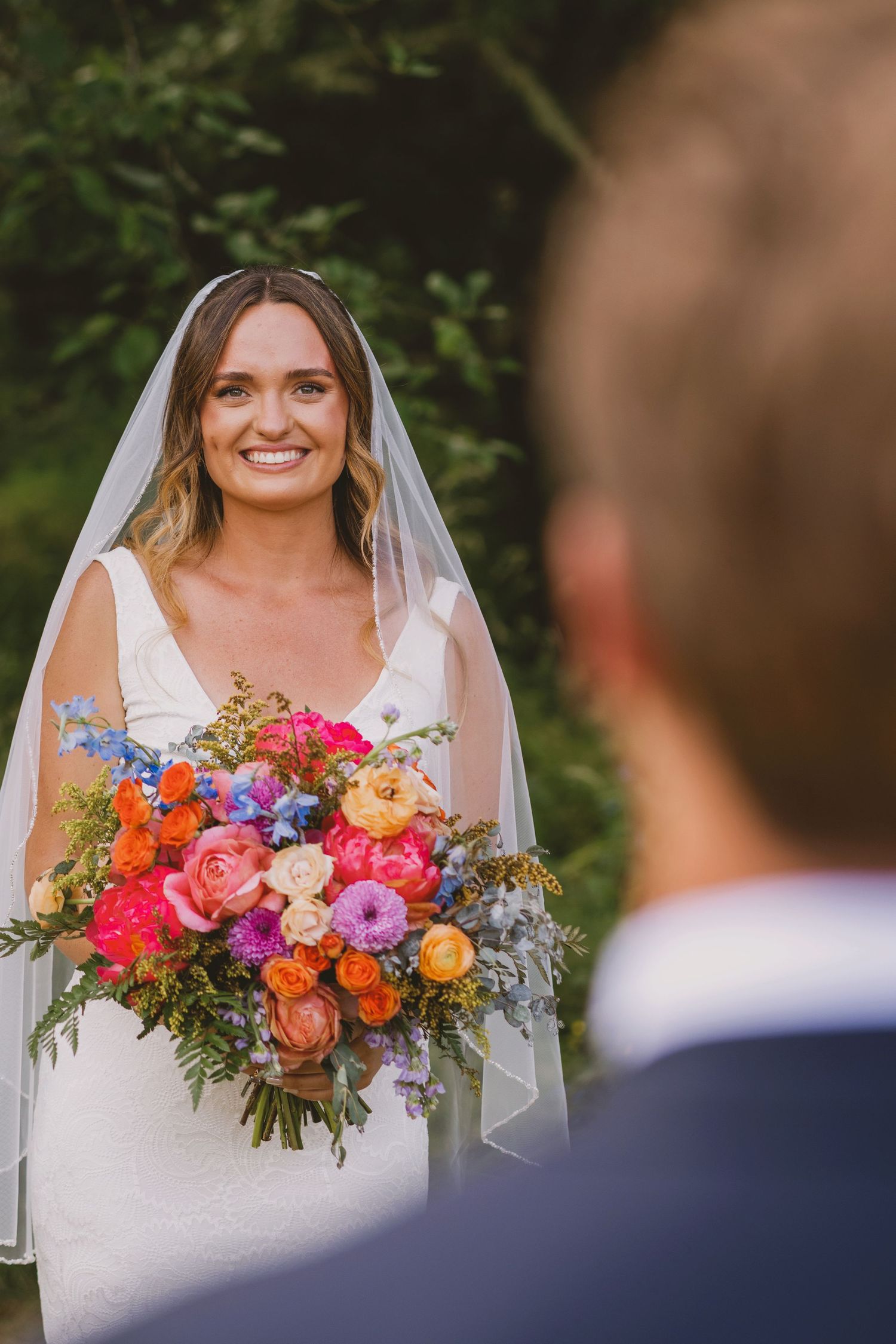 Smiling bride in white gown and veil holds vibrant colorful bouquet of pink, orange, and blue flowers outdoors.