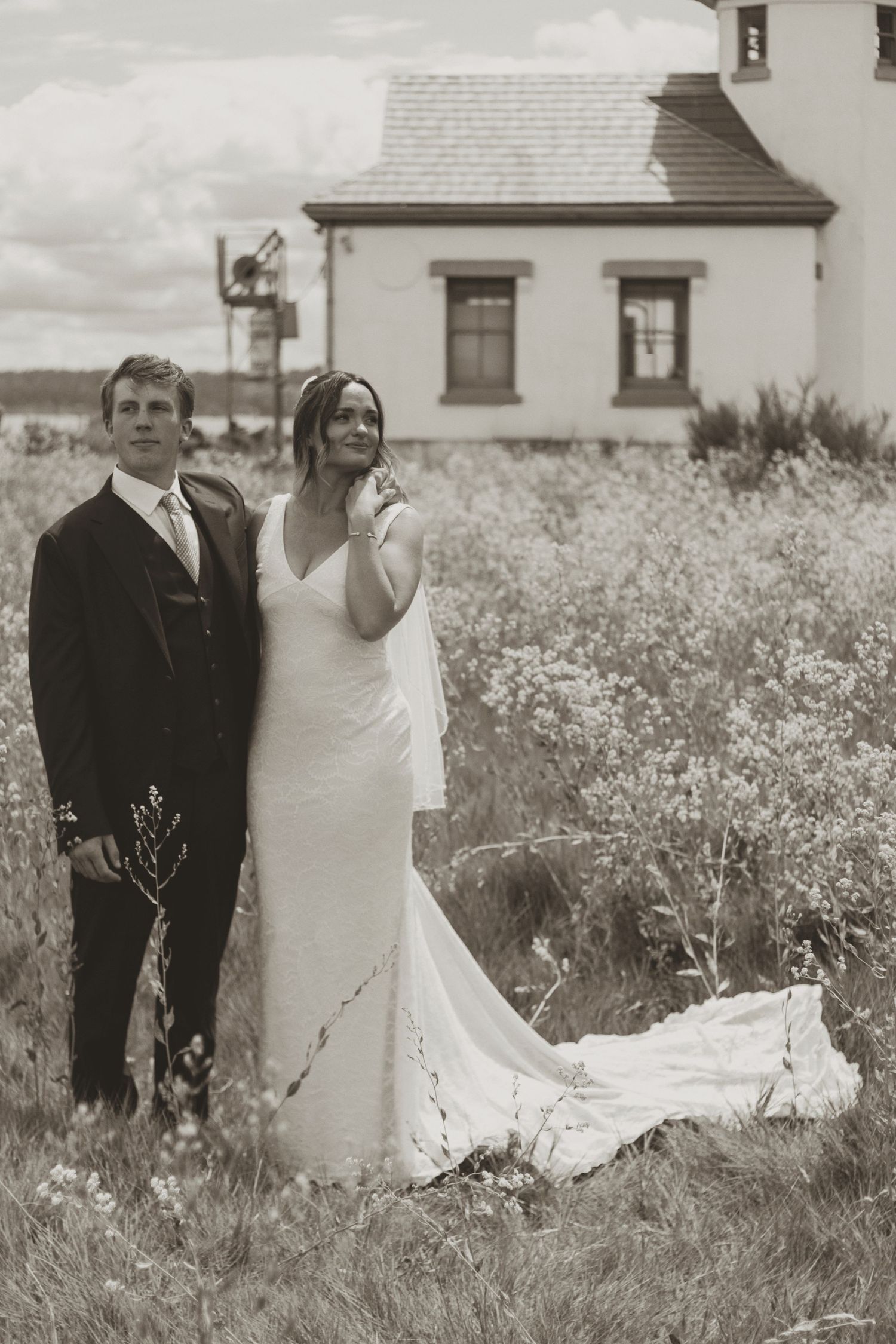 Bride and groom pose in a wildflower field beside a white country house in a romantic sepia-toned wedding photo.