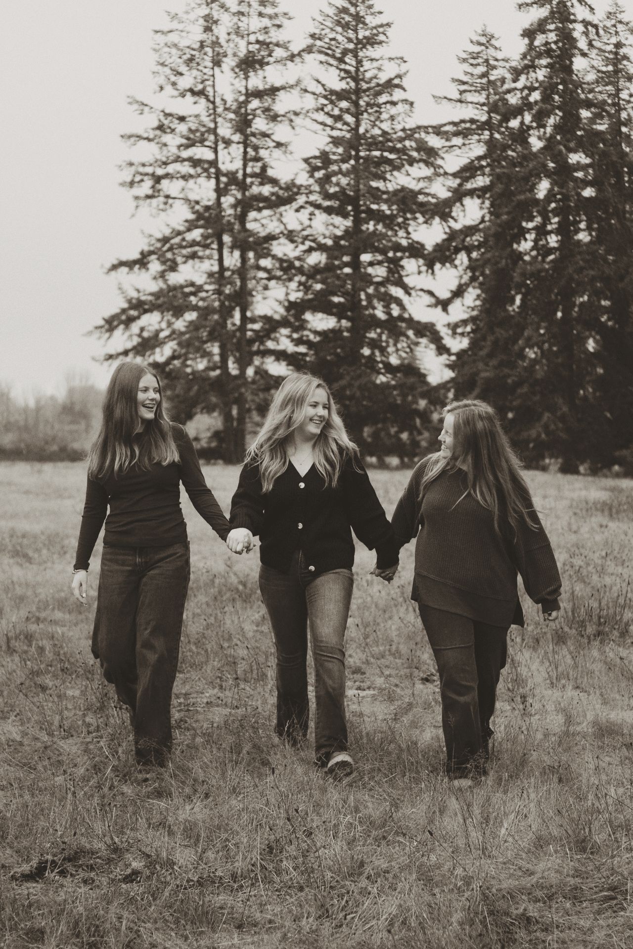 Black and white photo of three people walking through a grassy field holding hands with tall evergreen trees in background.