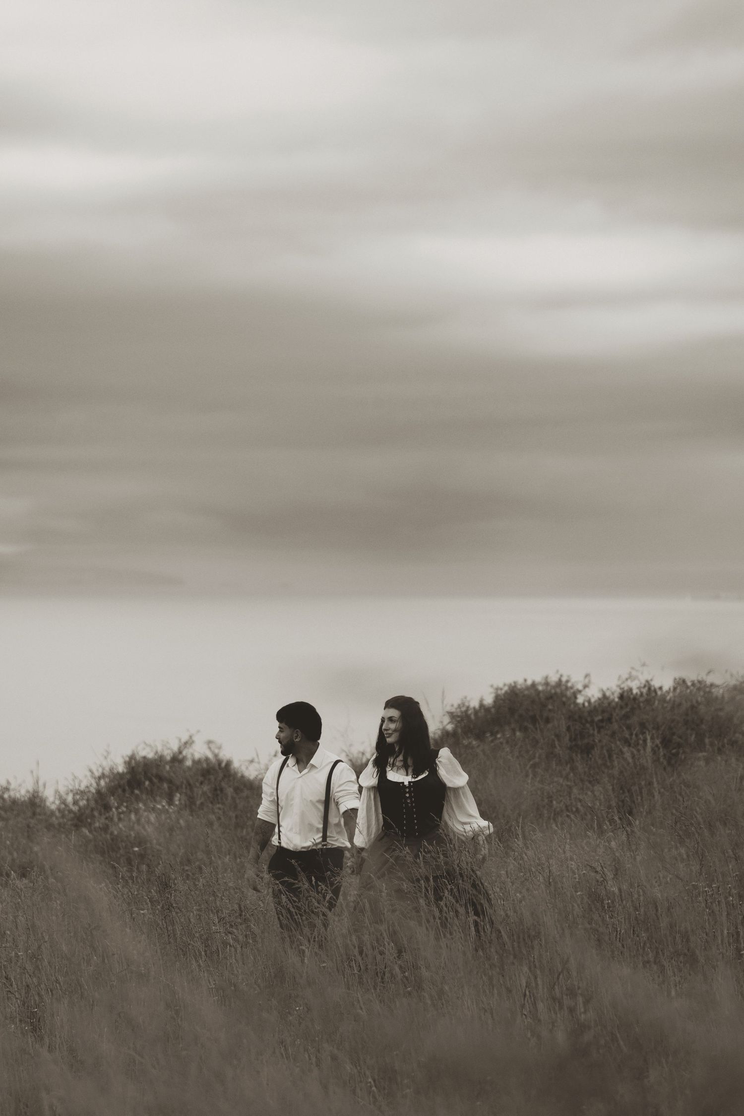Sepia-toned photo of a couple walking through tall grass fields under dramatic cloudy skies.
