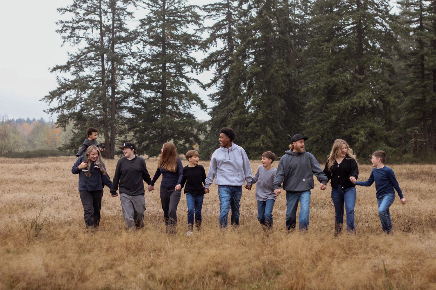 Group of people walking together holding hands in a line across a brown grassy field with evergreen trees in background.