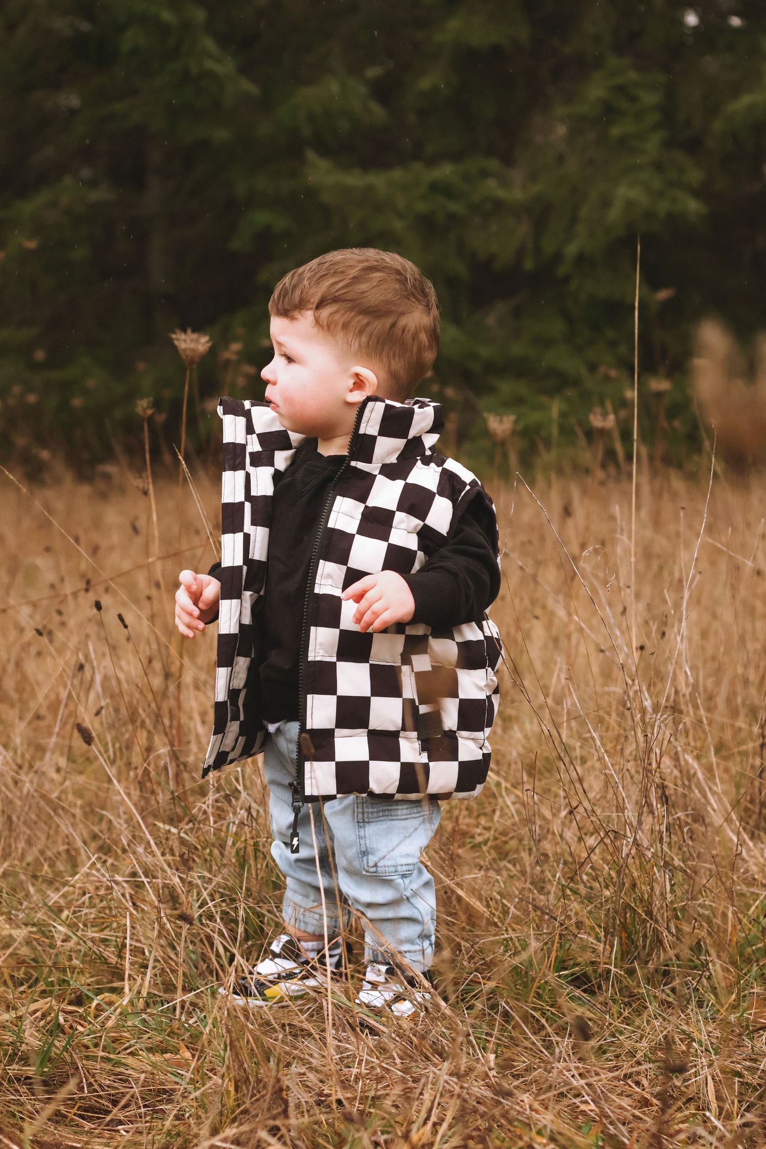 A toddler wearing a checkered sweater and jeans explores tall brown grass in an outdoor field.