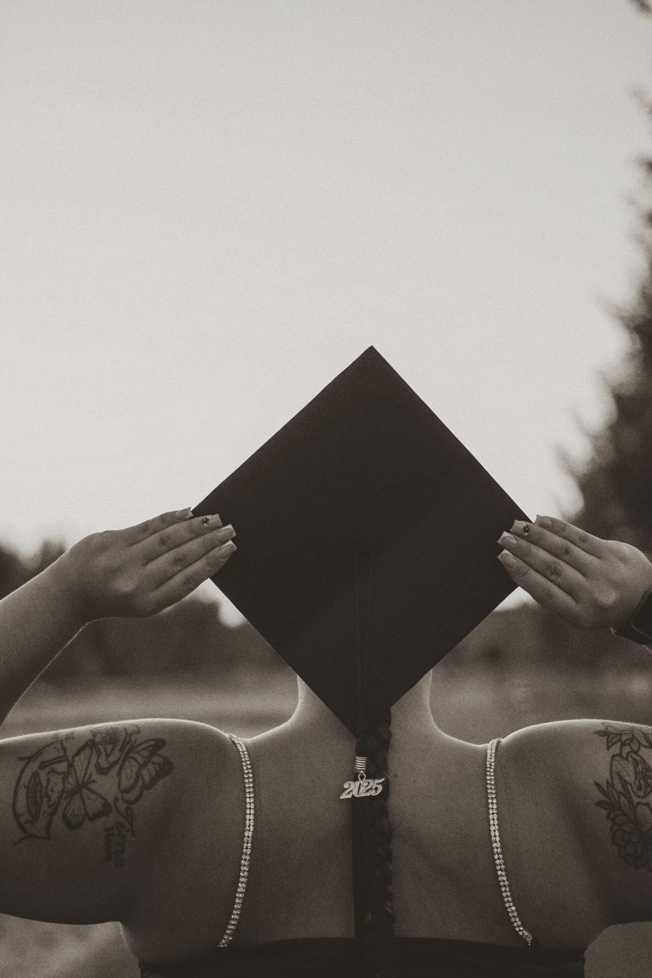 Black graduation cap held against cloudy sky in black and white photo.