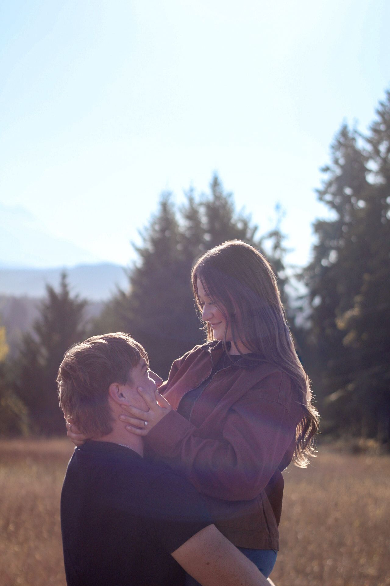 A couple embraces in a sunlit field with evergreen trees in the background.