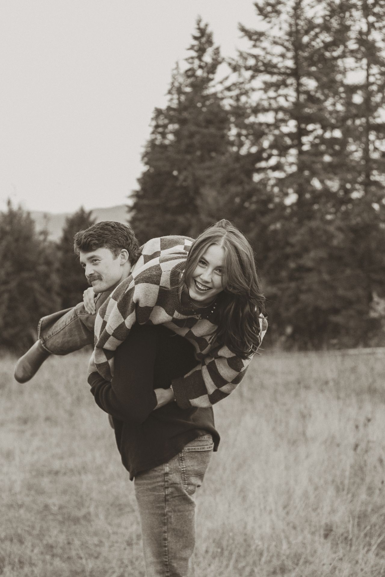 A couple in striped sweaters share a playful piggyback ride moment in a field with evergreen trees in the background.