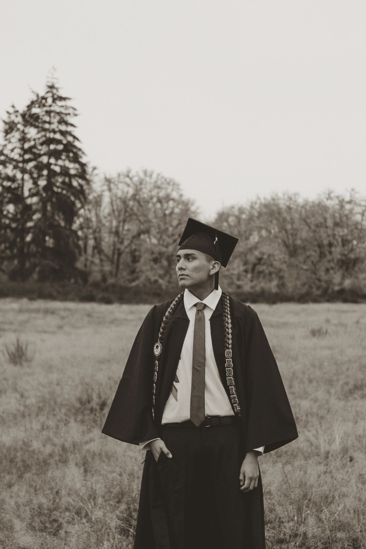 Black and white graduation photo showing graduate in traditional cap and gown standing in an outdoor field setting.