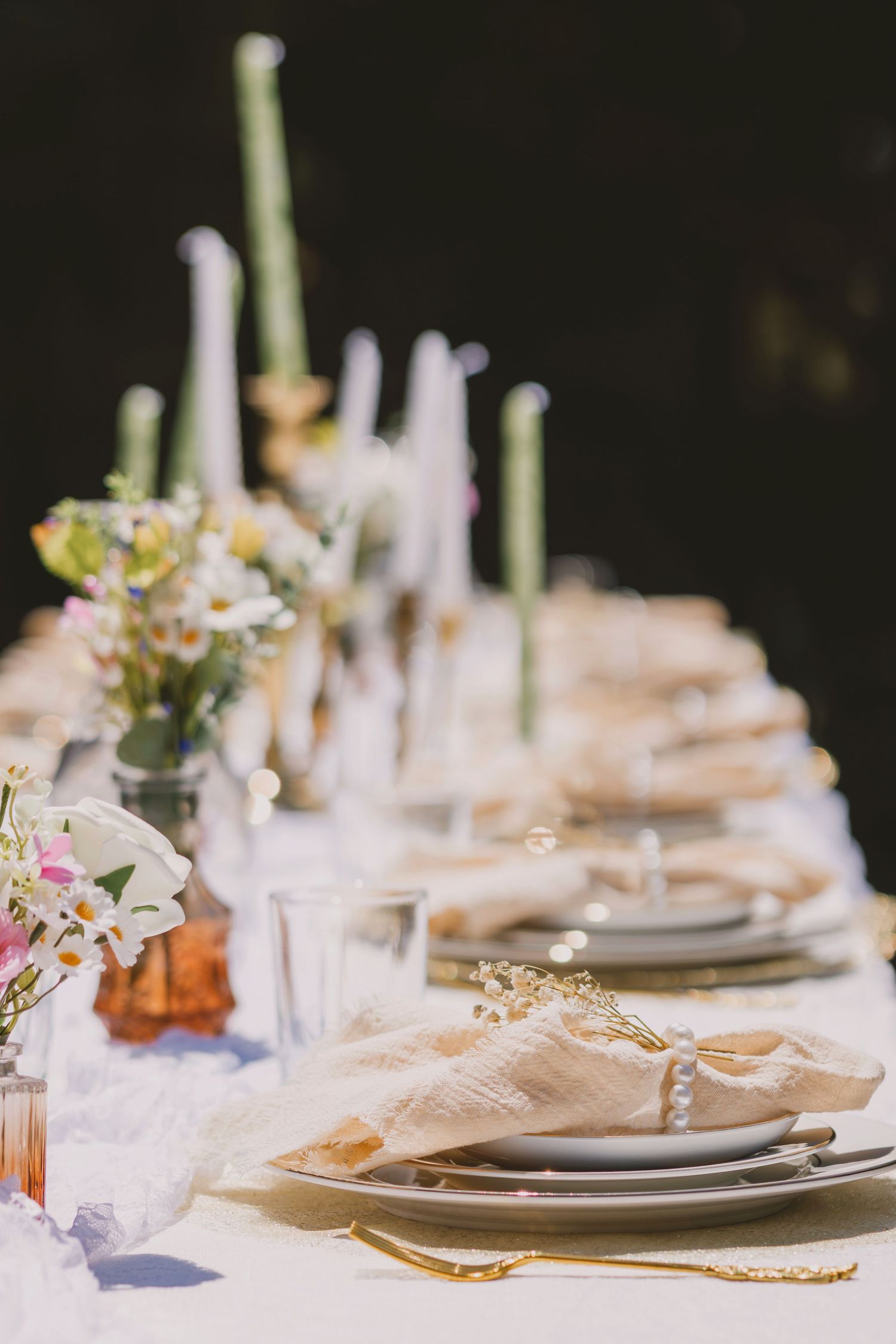 Elegant outdoor wedding table setting with green candles, floral arrangements, gold cutlery, and linen napkins on white tablecloth.