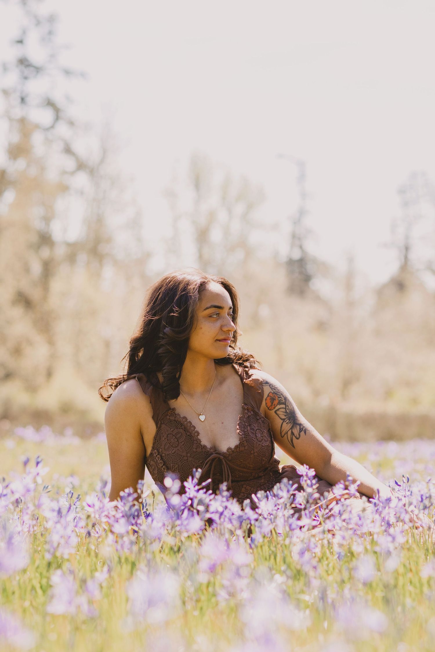 Woman in brown dress sitting among purple wildflowers in a sunlit meadow with trees in background.