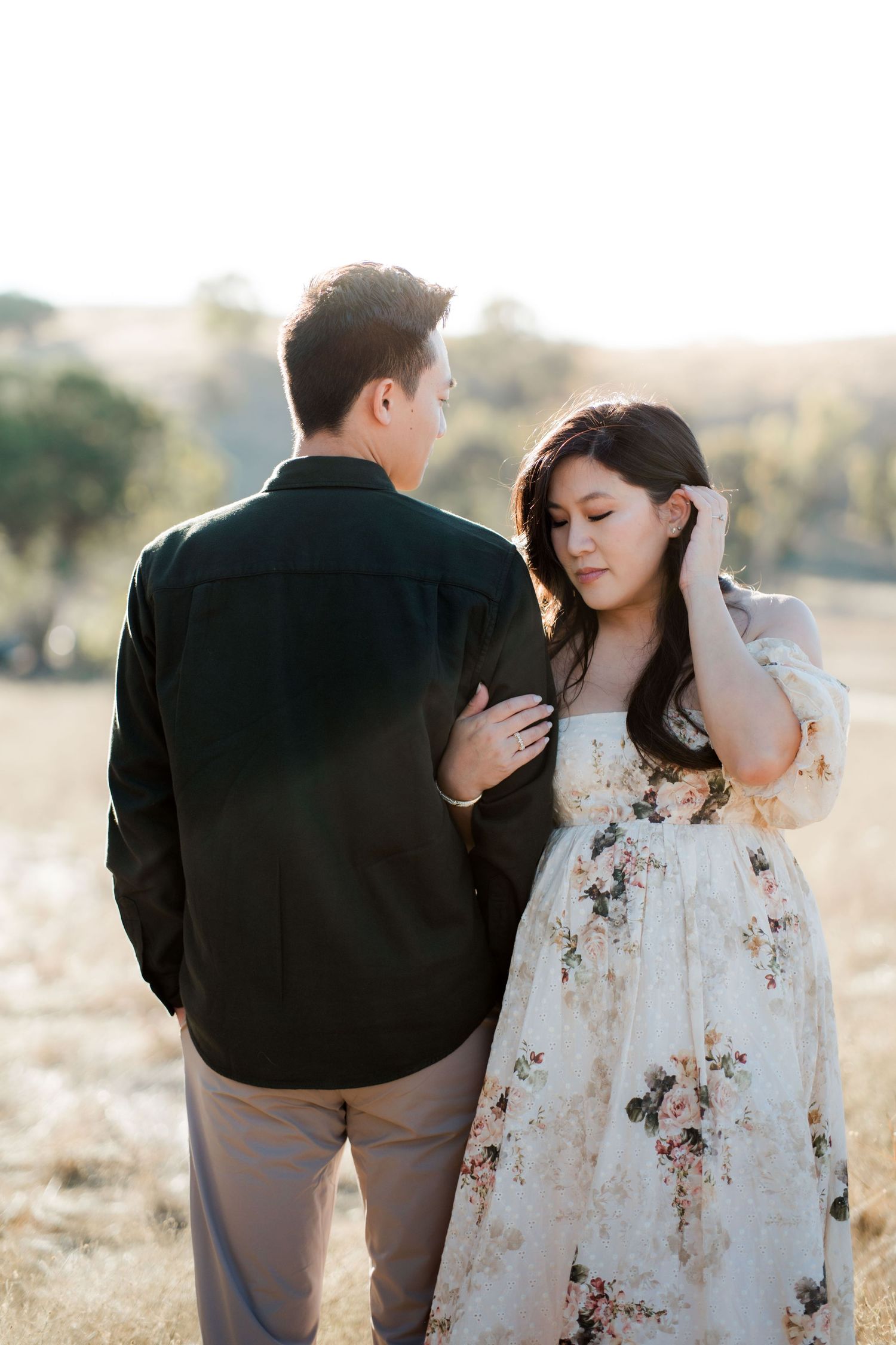 Couple in golden field at sunset, woman in floral dress holding mans arm in romantic pose.