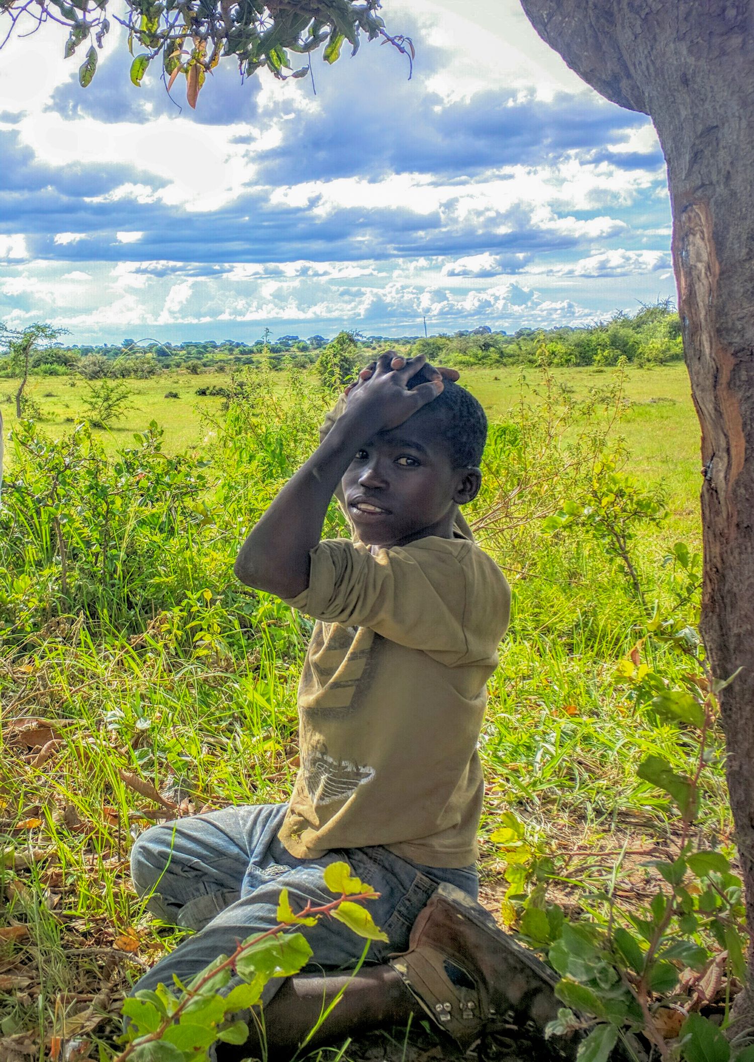 Young person sitting under tree in lush African savanna with bright blue sky and puffy clouds.