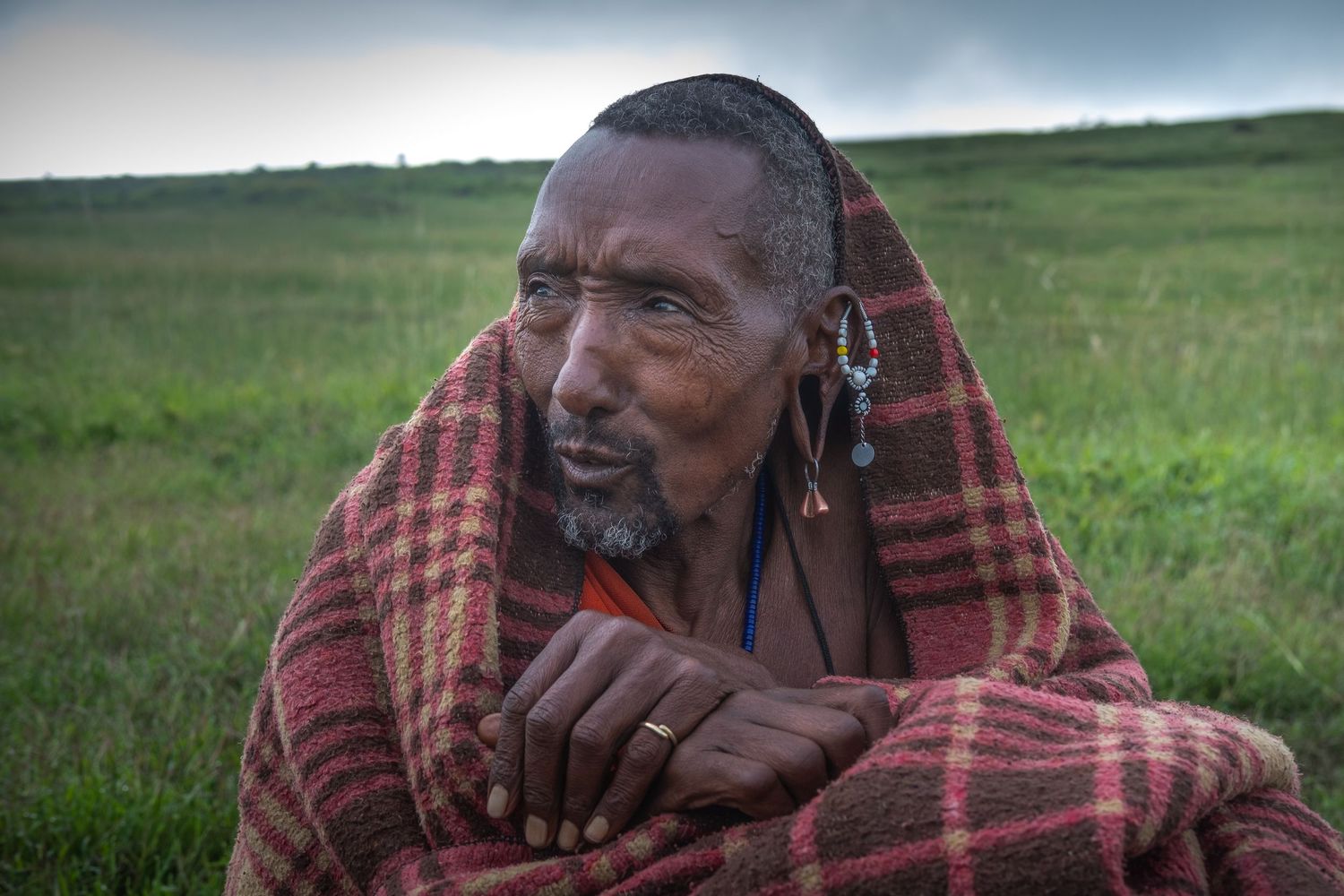 Person wearing traditional plaid clothing sitting in grassy savanna landscape.