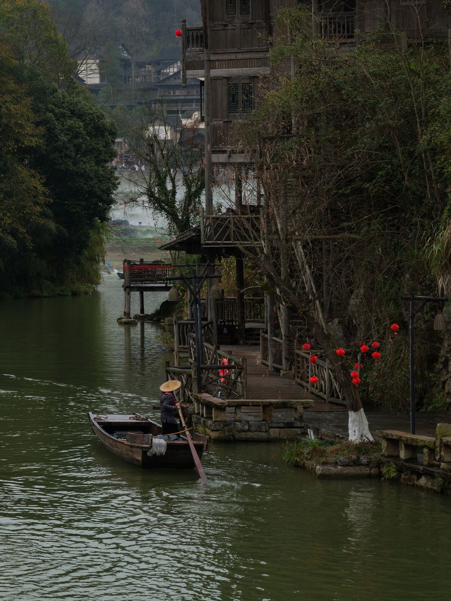 Traditional wooden houses on stilts along a river with red lanterns and small boats in the water.
