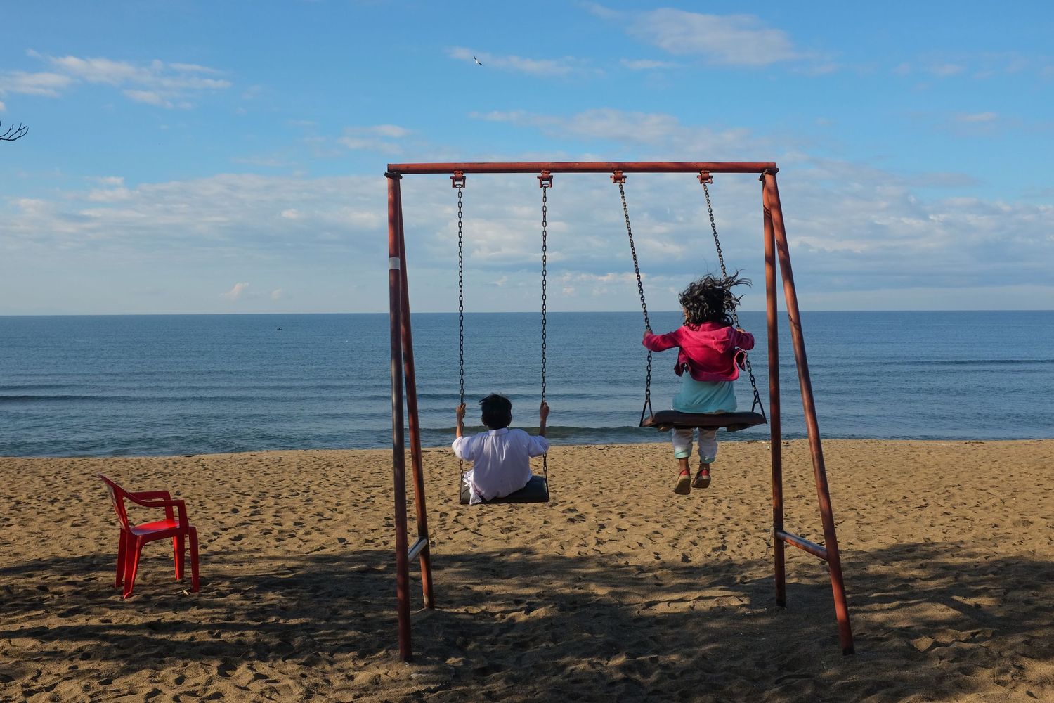 Red swing set on a sandy beach with ocean waves and blue sky in the background.