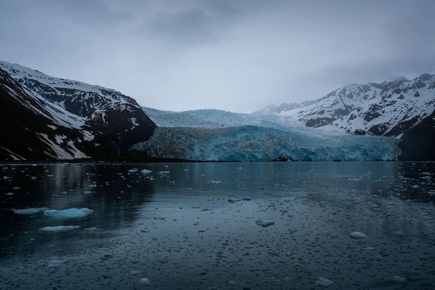 Snow-covered mountains reflect in a calm glacial lake under an overcast sky.